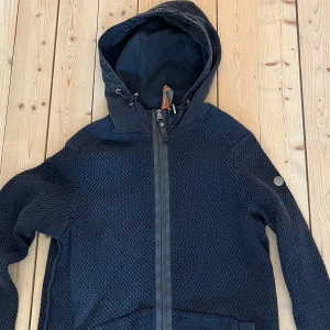 Mörkblå stickad jacka med huva - Mörkblå stickad cardigan med huva och dragkedja framtill. Jackan har lång ärm och en diskret patch på ena ärmen. Perfekt för kyligare dagar och har en sportig men stilren look. Sökord: cardigan, grisch 