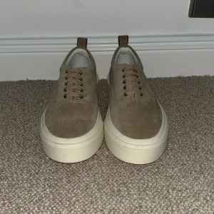 Beige sneakers från Eytys med tjock sula - Säljer ett par beige sneakers från Eytys med kraftig vit plattformssula och snörning. Skorna är i mocka och har en minimalistisk design med lila detalj baktill. Perfekta för dig som gillar stilrena och trendiga skor.