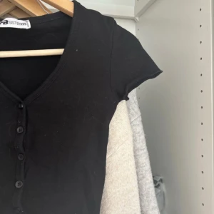 Svart croppad topp med knappar från new yorker - Svart croppad topp från new Yorker med korta ärmar och v-ringning i storlek xxs. Toppen har knappar framtill och en lätt ribbad struktur. Perfekt att styla med jeans eller kjol och aldrig använd. 💕
