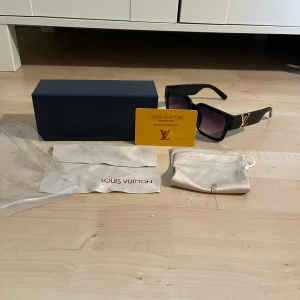 Svarta solglasögon från - Stilrena svarta solglasögonmed fyrkantig form och breda skalmar. På skalmarna finns en stor guldfärgad logga. Glasen är mörka med en lätt toning. Kommer med originalfodral, påse och certifikat. Oanvända, pris kan diskuteras