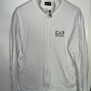 Vit zip-jacka från EA7 Emporio Armani - Säljer en stilren vit jacka från EA7 Emporio Armani med dragkedja framtill och logga på bröstet. Jackan har hög krage och långa ärmar. Perfekt för en clean och sportig look.