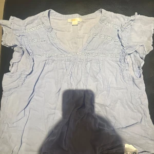 Ljusblå blus med volangärmar - Söt ljusblå blus med volangdetaljer på ärmarna och spets längs bröstet. Blusen har en lös passform och är tillverkad i ett lätt material, perfekt för varmare dagar.