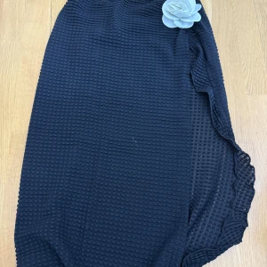 Svart långkjol med volang och blomdetalj - Svart långkjol i strukturerat tyg med volang längs ena sidan och en stor dekorativ blomma upptill. Kjolen har en elegant och unik look, perfekt för dig som vill sticka ut lite extra.