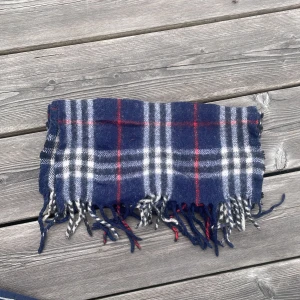 Blårutig halsduk från Burberry - Klassisk blårutig halsduk från Burberry i 100% lammull. Halsduken är i sådär skick, den är tvättad och då alltså lite hårdare än den ska vara den har några hål mm. Självklart äkta