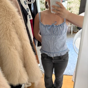 Blåvit randig linnetopp med rosett - Säljer en söt blåvit randig linnetopp med smala axelband och volangkant nertill. Toppen har en liten rosett framtill och är perfekt för varma dagar.