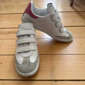 Vita Beth sneakers med kardborre från Isabel Marant - Säljer ett par vita sneakers från Isabel Marant med grå mockadetaljer och rosa hälparti. Skorna har tre breda kardborreband och perforerade detaljer framtill. Märkeslogga i guld på hälen. Perfekta för en stilren och sportig look. De är något små i storleken, skulle säga att de passar en storlek 40 också! Kan mötas upp i innerstan eller skicka. 
