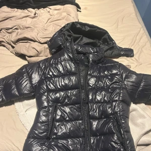 Svart pufferjacka från lager - Säljer en svart pufferjacka med huva och dragkedja framtill. Jackan har en glansig yta och två fickor med dragkedja. Perfekt för kyligare dagar och enkel att matcha med olika outfits. Denna är i storlek M och är knappt använd!