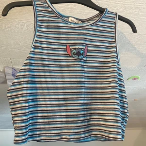 Randig ribbad linnetopp från Disney - Säljer en ribbad linnetopp från Disney i blått, vitt och svart med broderad Stitch på bröstet. Toppen är ärmlös och har ett randigt mönster. Perfekt för dig som gillar Disney och vill ha en färgglad och bekväm topp.