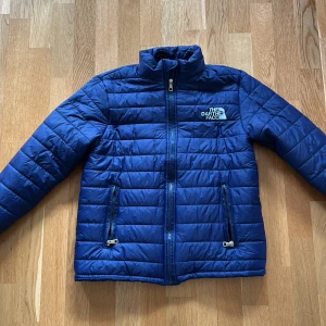 Blå pufferjacket med luva - Blå lättare pufferjacka. Jackan har en avtagbar huva i grått tyg. 