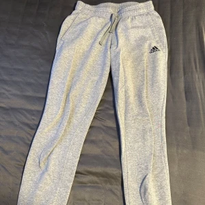 Grå mjukisbyxor från Adidas - Grå mjukisbyxor från Adidas med resår och snörning i midjan samt muddar vid bensluten. Byxorna har Adidas-logga på vänster lår och är tillverkade i mjuk bomullsblandning. Perfekta för en avslappnad stil. De här oanvända.
