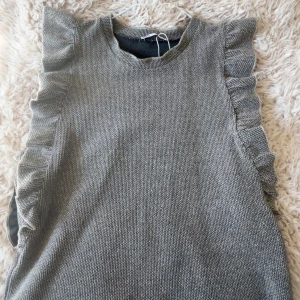 Grå stickad topp med volang från Zara - Supersnygg grå stickad topp från Zara med volangdetaljer längs sidorna. Rund halsringning och korta ärmar ger en cool och trendig look. Perfekt att styla med jeans eller kjol!