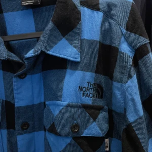 Blå och svart rutig skjorta från The North Face - Säljer en blå och svart rutig skjorta från The North Face. Skjortan har klassisk krage, knäppning framtill och två bröstfickor med knappar. Perfekt för dig som gillar en avslappnad och stilren look.