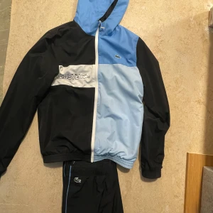 Svart och ljusblå vindjacka från Lacoste, svarta byxor  - Snygg vindjacka från Lacoste i svart och ljusblått med vit dragkedja. Svarta byxor som följer med dressen. Bra skick. 