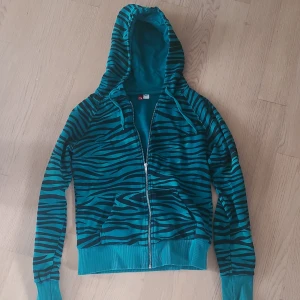 Turkos hoodie med zebramönster - Säljer en turkos hoodie med svart zebramönster och dragkedja framtill. Tröjan har huva, långa ärmar och ribbade muddar vid ärmslut och nederkant. Kom privat för ytterligare frågor!