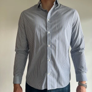 Blåvit randig skjorta från H&M - Säljer en stilren blåvit randig skjorta från H&M i slim fit-modell. Skjortan har långa ärmar, klassisk krage och knäppning framtill. Perfekt för dig som gillar en enkel men snygg look.