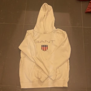 Vit hoodie från GANT med broderad logga - Vit hoodie från GANT med broderad logga och sköld på bröstet. Klassisk känguruficka framtill och huva med dragsko. Perfekt för en avslappnad stil. Slitningar som man ser på andra bilden, annars bra skick. Priset kan diskuteras! 