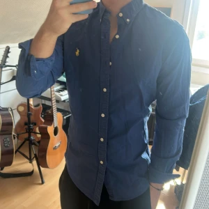 Blå skjorta från Ralph Lauren - Säljer en klassisk blå skjorta från Ralph Lauren med gul broderad logga på bröstet. Skjortan har knappar hela vägen, button-down krage och lång ärm. Perfekt för dig som gillar stilrena plagg med snygga detaljer.