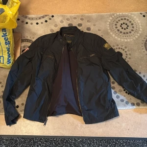 Svart vindjacka från G-Star RAW - Säljer en svart vindjacka från G-Star RAW med dragkedja framtill och två bröstfickor med dragkedja. Jackan har lång ärm och patchdetaljer på axeln och ärmen. Perfekt för dig som gillar en sportig och stilren look.