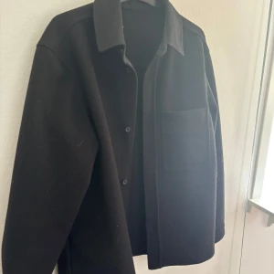 Svart overshirt med krage - Säljer en svart overshirt med klassisk krage och knäppning framtill. Jackan har en stor bröstficka och raka ärmar, perfekt för lager-på-lager-stil.