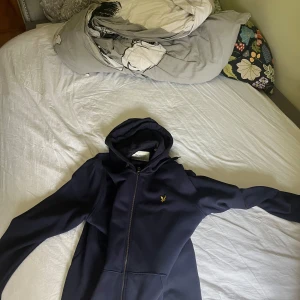 Mörkblå hoodie från Lyle & Scott - Säljer en mörkblå hoodie från Lyle & Scott med dragkedja och huva. Tröjan har en liten gul logga på bröstet och är tillverkad i mjukt material. Perfekt för en avslappnad stil. Pris kan diskuteras gällande snabbaffär bara använd 2 gånger.
