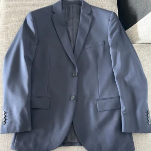 Gagliardi mörkblå kavaj med klassisk passform - Snygg mörkblå kavaj från Gagliardi med två knappar framtill och tre knappar vid ärmsluten. Klassisk krage och flera fickor både på utsidan och insidan. Perfekt för bal eller fest. Tunn fodring på insidan för extra komfort.