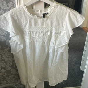 Vit blus med volang från Vero Moda - Säljer en söt vit blus från Vero Moda med volangdetaljer på ärmarna och broderat mönster. Blusen har korta ärmar, rund halsringning och knapp i nacken. Perfekt för vår och sommar!