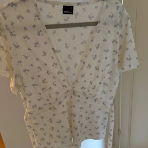 Vit blommig topp från Gina Tricot - Söt vit omlottopp från Gina Tricot med små blålila blommor. Toppen har korta ärmar, knytband i midjan och v-ringning framtill. Perfekt för vår och sommar, lätt och luftig i bomullsmaterial. Använd ett fåtal gånger, inköpt förra sommaren.