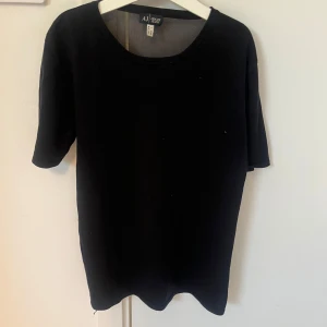 Svart t-shirt från Armani Jeans - Svart t-shirt från Armani Jeans med diskret logga i paljetter över ryggen. Klassisk rund halsringning och korta ärmar. Perfekt för en stilren och enkel look.