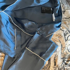 Blå pyjamasskjorta från H&M - Mysig blå pyjamasskjorta från H&M med vita kontrastkanter och bröstficka. Skjortan har lång ärm och klassisk krage, tillhörande byxor kan man även köpa till, perfekt för avslappnade kvällar hemma. Skjorta 75kr byxor 90kr båda för 155