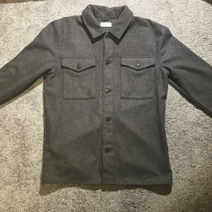 Grå overshirt  These Glory Days - Snygg grå overshirt från These Glory Days i storlek medium. Jackan har en klassisk krage och knäppning framtill med knappar. Två bröstfickor med lock och knappar ger en stilren look. Perfekt för lager-på-lager. Gjord av ull. 