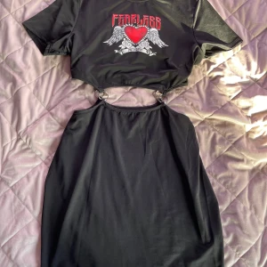 Svart cool alternative klänning med tryck - Cool svart alternativ klänning med ett tryck av ett rött hjärta och vingar samt texten 'FEARLESS'. Klänningen har korta ärmar och en unik design med öppna partier och ringdetaljer i midjan. Perfekt för en edgy look! Använd bara några några gånger! Stretchigt material! Öppen för prisförslag! 