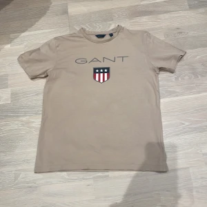 Beige t-shirt från GANT - Säljer en stilren beige t-shirt från GANT med ett tryck av deras logotyp och ett sköldemblem på framsidan. T-shirten är kortärmad och tillverkad i mjuk bomull, perfekt för en avslappnad stil.