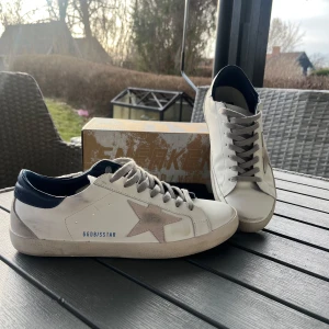 Golden Goose Super-Star sneakers - Snygga Golden Goose Super-Star sneakers i vitt med en ikonisk stjärndetalj på sidan. Skorna har grå snörning och en mörkblå häl. Perfekta för en stilren look. Priset är ej hugget i sten, och är öppen förhandling. Minsta fråga? Fråga på då ;)