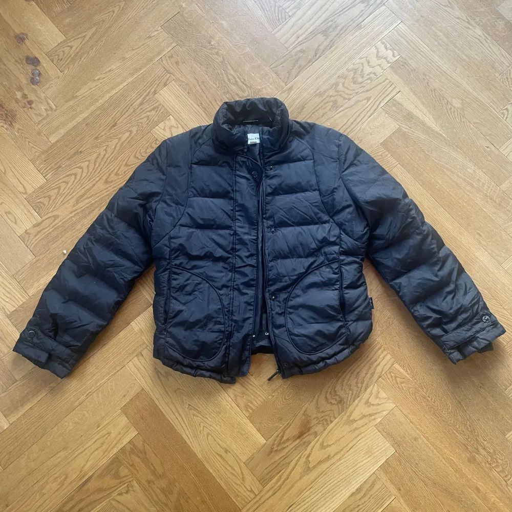 Säljer en svart pufferjacka från Calvin Klein. Jackan har en dragkedja framtill och långa ärmar. Har både dragkedja och knappar, samt som gummiband nertill så man kan spänna åt hur den sitter till sin egen preferens. Den är quiltad och har en stilren design med rundad krage. Perfekt för kyligare dagar. (På vissa knappar så har den blivit lite missfärgning, de sista bilden) Pris kan absolut diskuteras!❣️ Själv köpt secondhand för 400kr❤️. Takit.