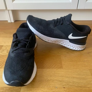 Svarta Nike sneakers - Säljer ett par svarta Nike sneakers med vit sula och klassisk Nike-logga på sidan. Skorna har en stickad ovandel och snörning för bra passform. Perfekta för både träning och vardag. Säljs i strlk 38,5