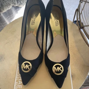 Svarta pumps från Michael Kors - Snygga svarta pumps från Michael Kors i mocka med spetsig tå och en elegant MK-logga framtill. Perfekta för att ge en stilren touch till din outfit. Storlek M-9 storlek 40, känns mer som 38,5 39. Klack 10 cm. 