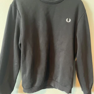 Svart sweatshirt från Fred Perry - Säljer en klassisk svart sweatshirt från Fred Perry med deras ikoniska lagerkrans-logga i vitt på bröstet och ryggen. Tröjan har långa ärmar och en rund halsringning. Perfekt för en stilren och avslappnad look.