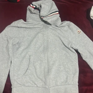 Grå huvtröja från Moncler - Snygg grå huvtröja från Moncler med dragkedja och ribbade muddar i rött, vitt och blått. Tröjan har en broderad Moncler-logga på ärmen och en bekväm passform. Perfekt för en stilren look.