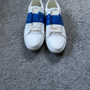 Valentino Garavani sneakers i vitt och blått - Snygga Valentino Garavani sneakers i vitt med en iögonfallande blå detalj över snörningen. Skorna har en klassisk design med rund tå och är tillverkade i skinn. Perfekta för att ge din outfit en lyxig touch.