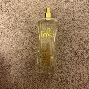 In Love Eau de Parfum från La Rive - Säljer en elegant flaska av parfymen 'In Love' från La Rive. Flaskan är genomskinlig med en gyllene spraypump och text i guld. Perfekt för den som vill ha en romantisk och sofistikerad doft.