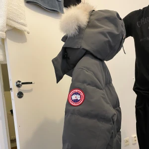 Grå Canada goose - Säljer en grå dunjacka från Canada Goose med en fluffig pälsdetalj på huvan. Jackan har en klassisk design med lång ärm och en praktisk dragkedja framtill. Perfekt för kalla vinterdagar. Storlek S