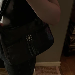 Svart axelväska med fransdetalj - Snygg svart axelväska i skinn med coola fransdetaljer och nitar på framsidan. Väskan har en justerbar axelrem och flera praktiska fack med dragkedja. Perfekt för att bära med sig det viktigaste med stil.