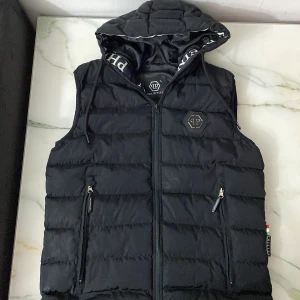 Svart dunväst från Philipp Plein - Snygg svart dunväst från Philipp Plein med huva och dragkedja. Västen har två fickor med dragkedjor och en broderad logotyp på bröstet. Perfekt för kyligare dagar.