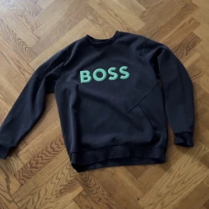 Svart sweatshirt från Boss - Säljer en mörkblå sweatshirt från Boss med grönt tryck på som är i nyskick. tröjan har  en klassisk rund halsringning. Perfekt för en avslappnad stil. storlek L