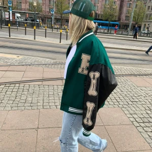 Grön och svart bomberjacka från Aelfric Eden - Snygg grön bomberjacka med svarta ärmar och vita detaljer från Aelfric Eden. Jackan har broderade bokstäver och mönster på ärmarna och bröstet. Perfekt för en cool och avslappnad stil.