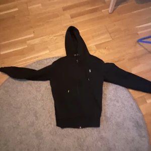Svart hoodie med dragkedja - Säljer en svart hoodie med dragkedja och huva. Den har långa ärmar och en enkel design. Perfekt för en avslappnad stil.
