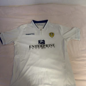 Vit fotbollströja från Macron - Säljer en vit fotbollströja från Macron med Leeds Uniteds emblem på bröstet. Tröjan har korta ärmar och en klassisk krage med blå detaljer. Den har tryck av Enterprise Insurance på framsidan och Help-Link Home på baksidan. Perfekt för fotbollsfans!