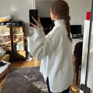 Vit stickad tröja - Säljer en mysig vit stickad tröja med hög krage och långa ärmar. Perfekt för kyliga dagar och ger en stilren look. Tröjan har en lös passform och ribbade detaljer vid ärmslut och nederkant.