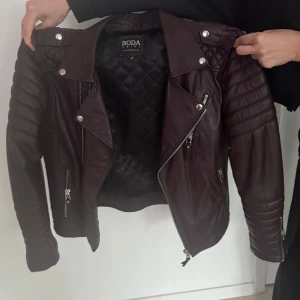 Vinröd skinnjacka från Boda Skins - Säljer en snygg vinröd skinnjacka från Boda Skins. Jackan har en quiltad insida och dragkedjor på framsidan och ärmarna. Den har en cool bikerstil med axelknappar och ribbade detaljer på ärmarna.