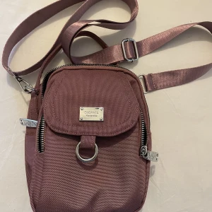 Axelremsväska  - En ny väska som aldrig är använd. Kan såklart användas hur man vill men tanken är att slänga tvärs över axeln. Du ser färgen bäst på sista bilden, Otroligt många smarta fack.  20 cm hög och 15 cm bred. Med band som går att göra längre eller kortare.